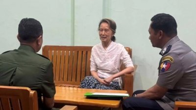 রাষ্ট্রীয় গণমাধ্যমে সু চির একটি ছবি প্রকাশ করা হয়েছে, যেখানে দেখা যায়, তিনি একটি কাঠের বেঞ্চে বসে আছেন এবং পাশে রয়েছেন ইউনিফর্ম পরা দুই নিরাপত্তাকর্মী। ছবি: এপি