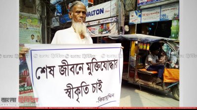 ব্যানার হাতে রাস্তায় দাঁড়ানো মুক্তিযোদ্ধা কিয়ামউদ্দীন 